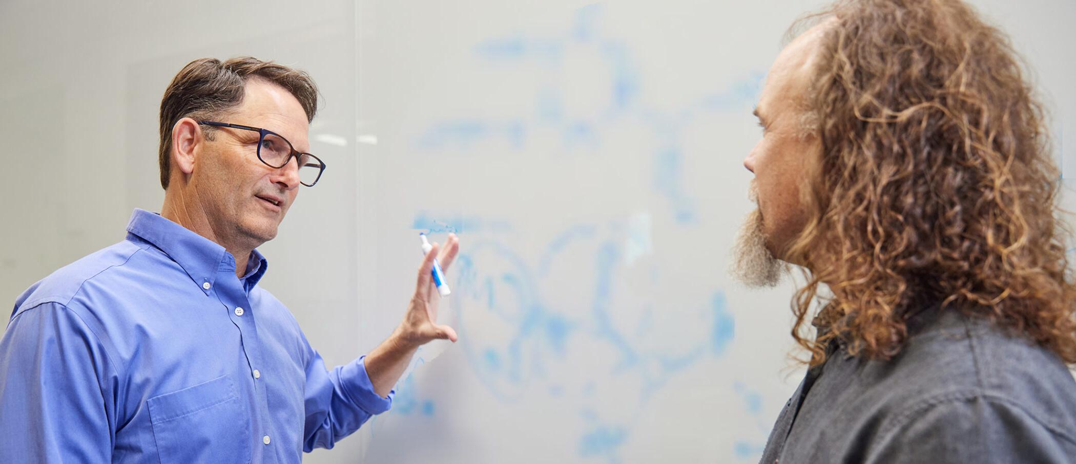 博彩平台网址大全 scientists Brett Crawford and Dan Wendt talking by a whiteboard with a scientific formula